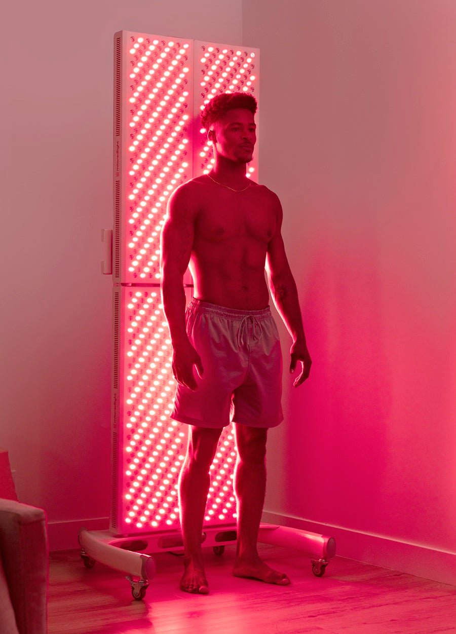 A shirtless man in shorts stands near a glowing MitoPRO+ | Red Light Therapy Panel Series v2 by Mito Red Light, bathing in red and near infrared light, in a sleek, modern room illuminated with a reddish-pink glow.