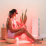 A woman in sunglasses sits on a cushion in front of the Mito Red Light MitoPRO+ | Red Light Therapy Panel Series v2, with a potted plant and small basket nearby in a minimalist room.
