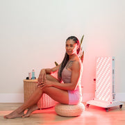 A woman with long braids sits on a round cushion in front of the Mito Red Light MitoPRO+ | Red Light Therapy Panel Series v2, smiling in athletic wear. Skincare products rest on a wicker basket beside her, with a plant in the background.