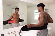 A shirtless man wearing gray shorts stands before a bathroom mirror, holding the MitoQUAD™ Wavelength Belt by Mito Red Light with red lights on his lower back, possibly using hands-free red light therapy for pain relief.