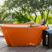 A person relaxes outdoors on a wooden deck in the Icebound Essentials Big Tex Endurance Plunge Tub, colored UT burnt orange, surrounded by Texas greenery and trees, with a black machine next to the tub.