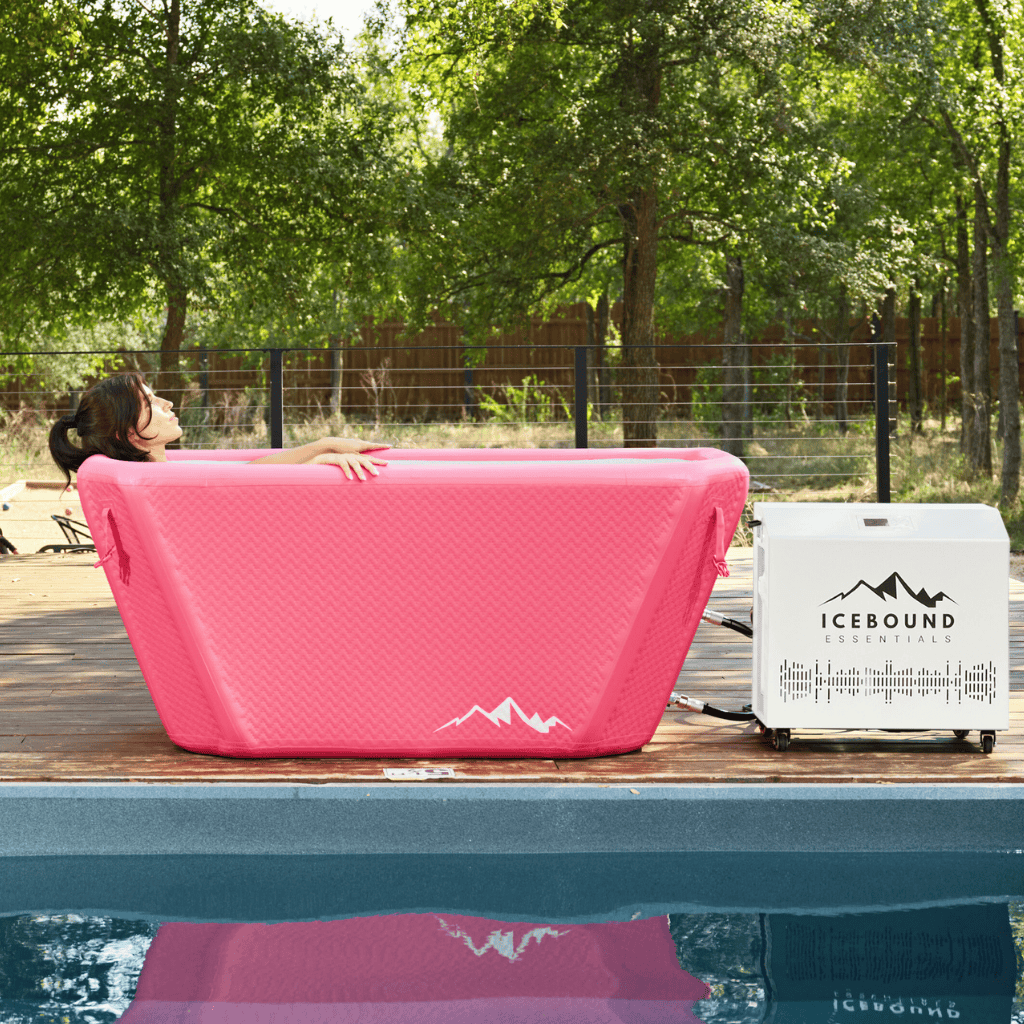 A person relaxes in an Icebound Essentials Pink Endurance Plunge + Chiller by the pool, with trees and a wooden fence in the background.