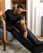 A man at home, seated on a gray chair and wearing a black T-shirt, zips up the HEALiX Infrared HEALiX Flow compression therapy leg sleeve over his right leg.