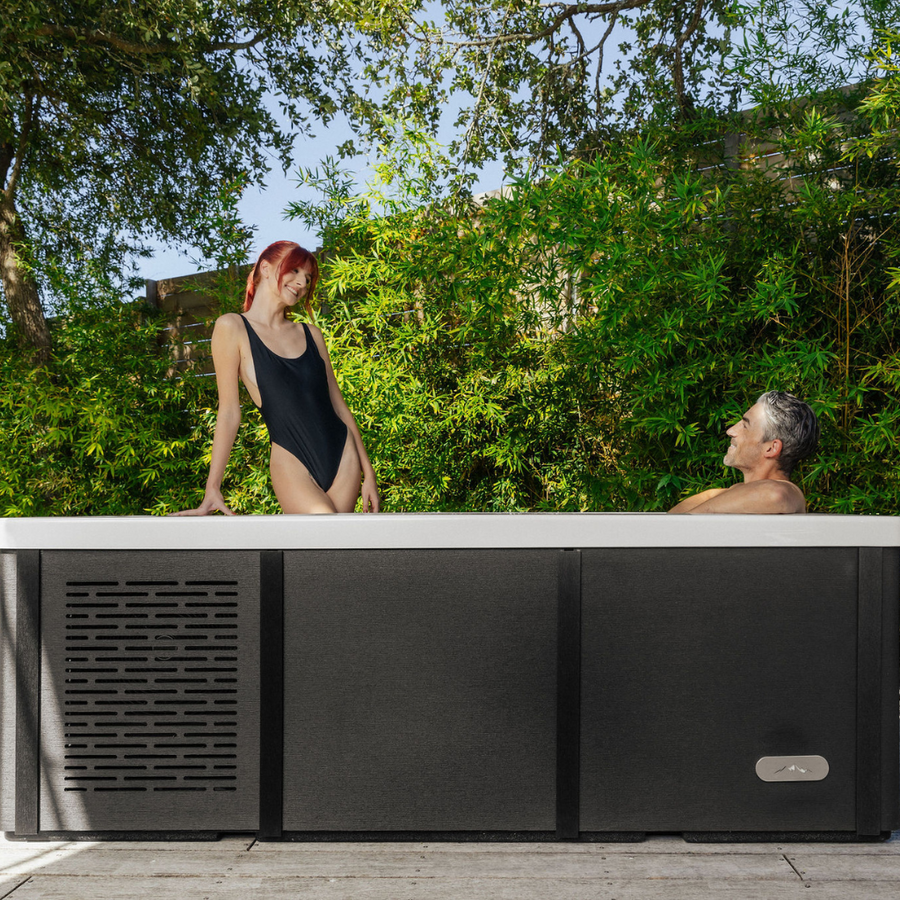 A woman in a black swimsuit sits on the edge of The Resolute Pro by Icebound Essentials, an outdoor hot tub with advanced Ozone sanitization, smiling at a man relaxing inside amid lush greenery on a sunny day.