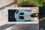 A woman in a black swimsuit relaxes in The Resolute Pro by Icebound Essentials, a modern outdoor professional-grade bathtub set on wooden decking amid greenery, seen from above.